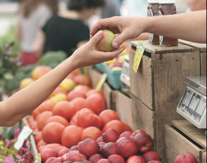 Immagine di due mani che si passano una mela e sullo sfondo una bancarella di mercato con della frutta e verdura